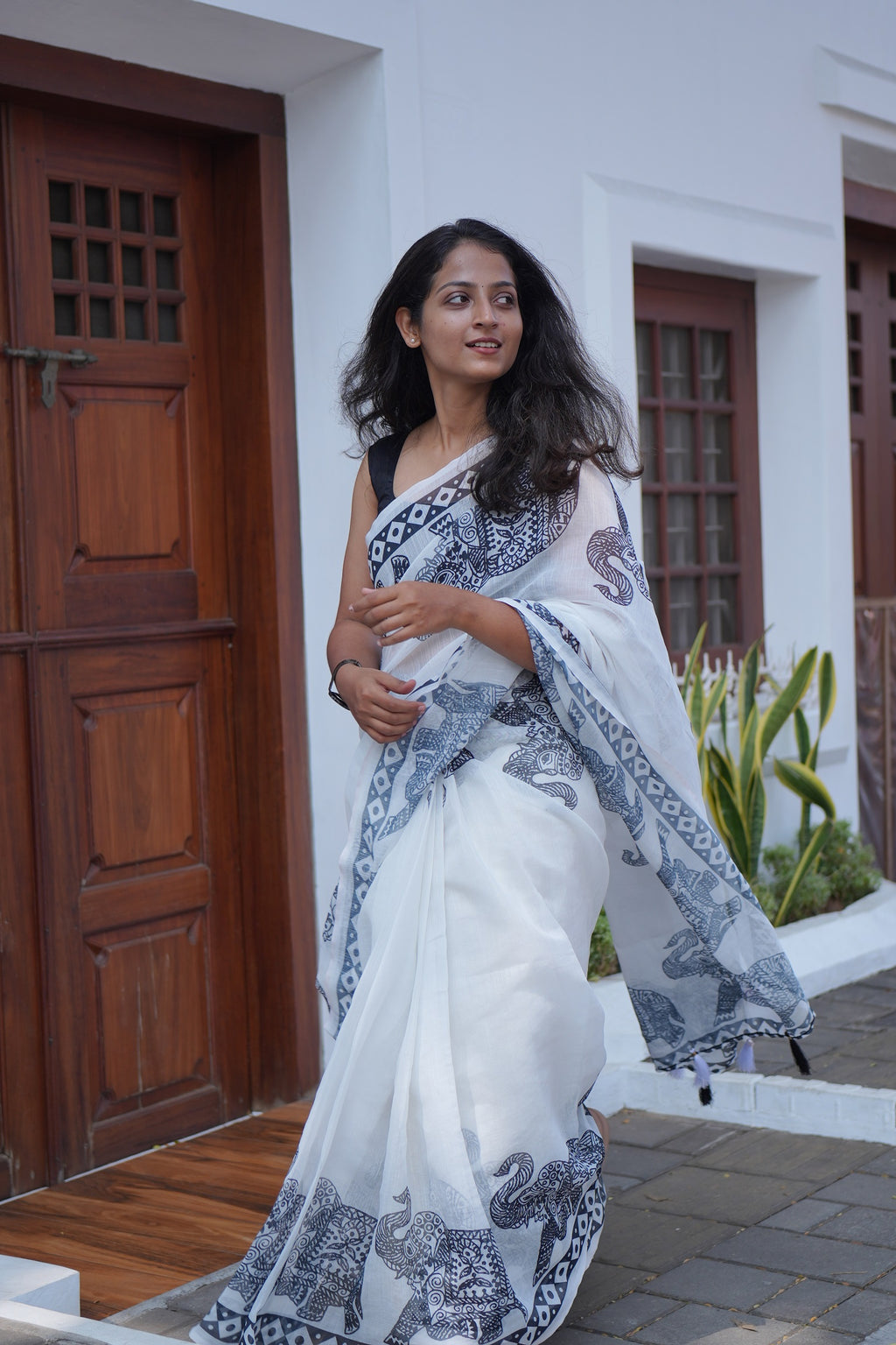 Woman in a white saree with blue patterns standing in front of a white building with wooden doors.